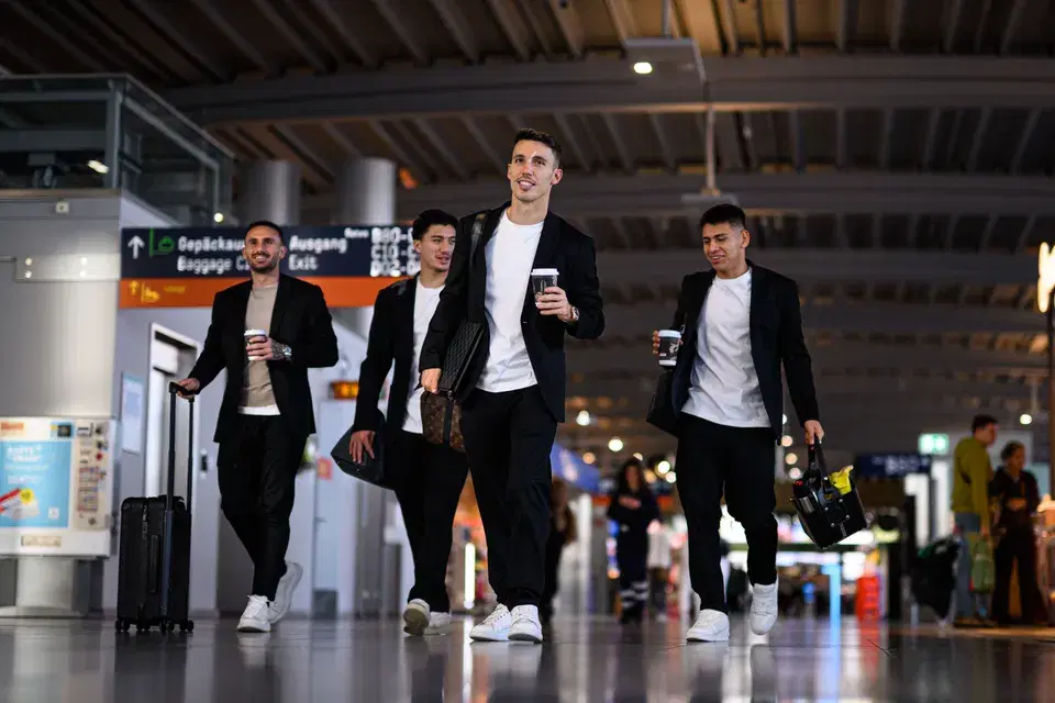 4 Fußballer in Sakkos laufen im Flughafen mit Tasche und Kaffee entlang